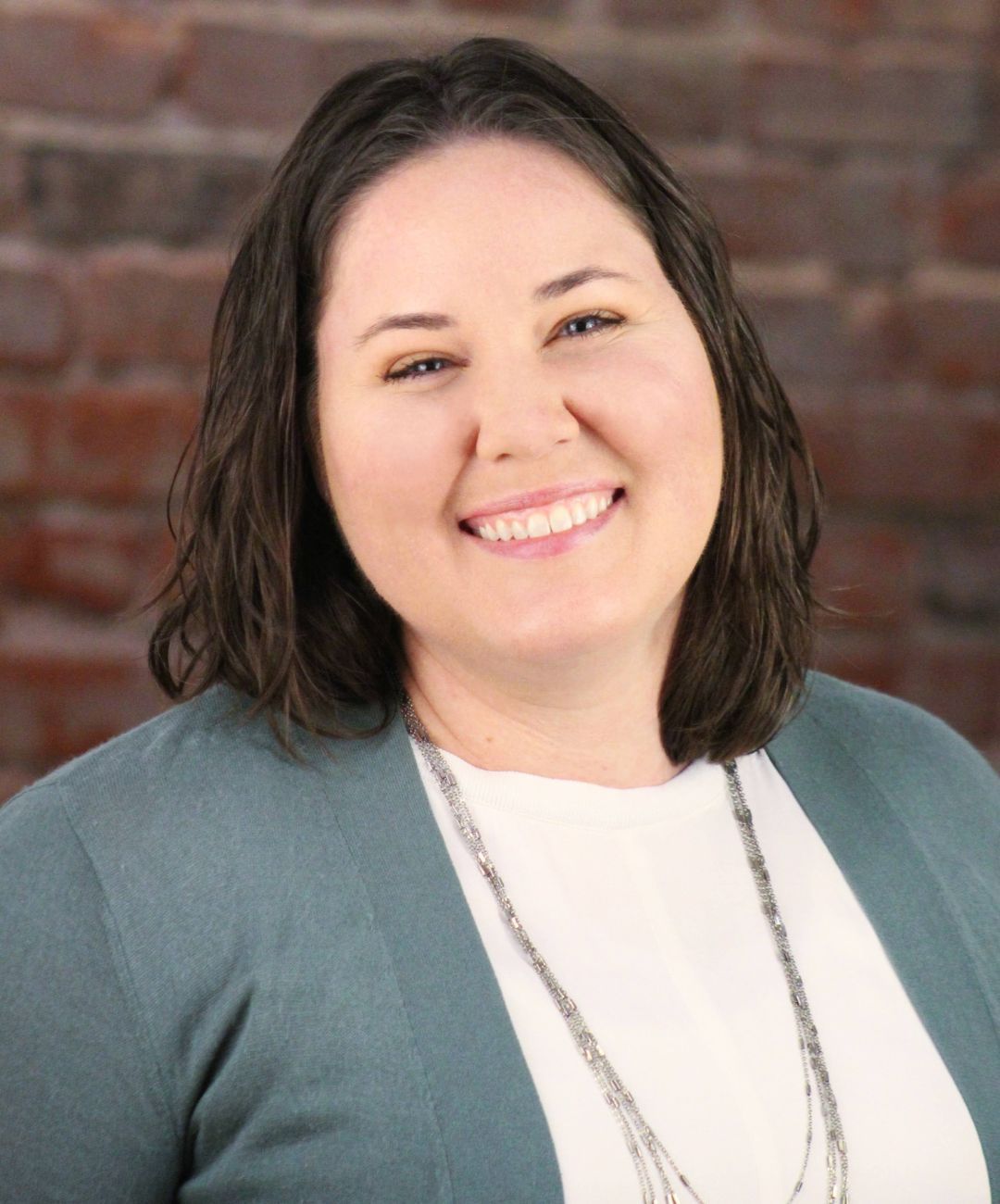 Smiling person in a teal cardigan over a white top, standing against a brick wall.