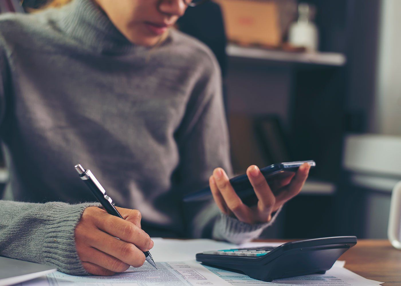 Person writing on paper while holding a smartphone beside a calculator on a desk.