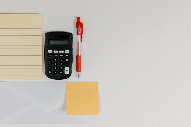 Notepad, calculator, and red pen beside a yellow sticky note on a white desk