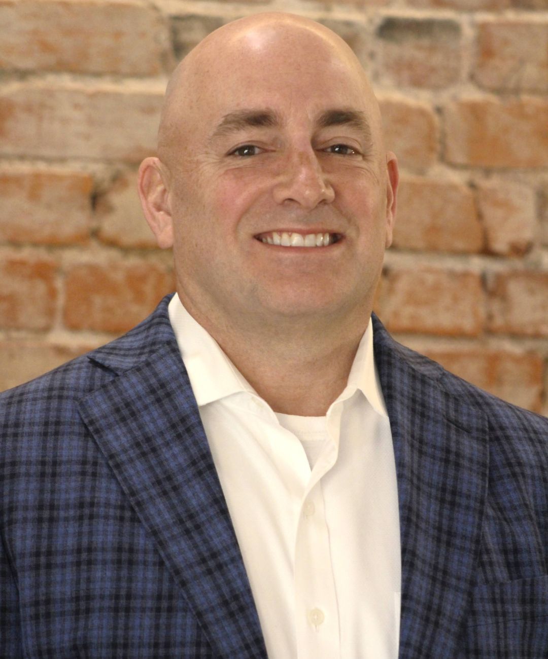 Smiling bald man in a blue plaid blazer and white shirt against a brick wall.