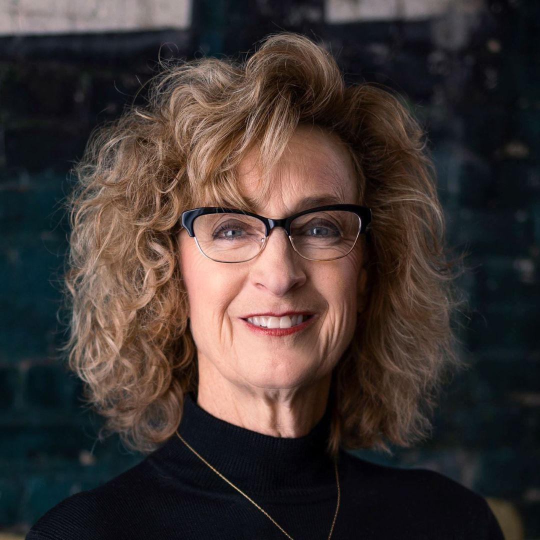Portrait of a smiling woman with curly blonde hair and glasses against a dark background