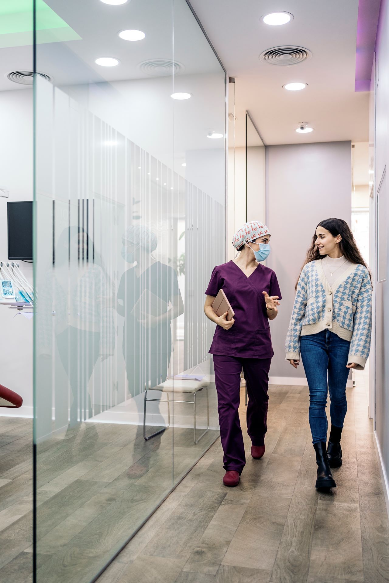 Two healthcare workers in purple scrubs walk and talk in a bright hospital hallway.