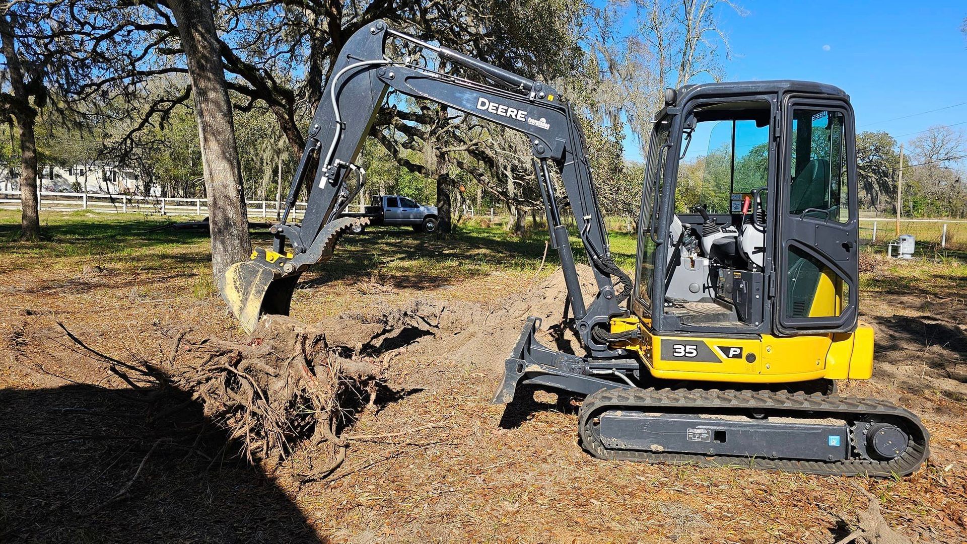 A yellow and gray mini excavator is positioned in a wooded area, digging into the earth near the base of a tree.