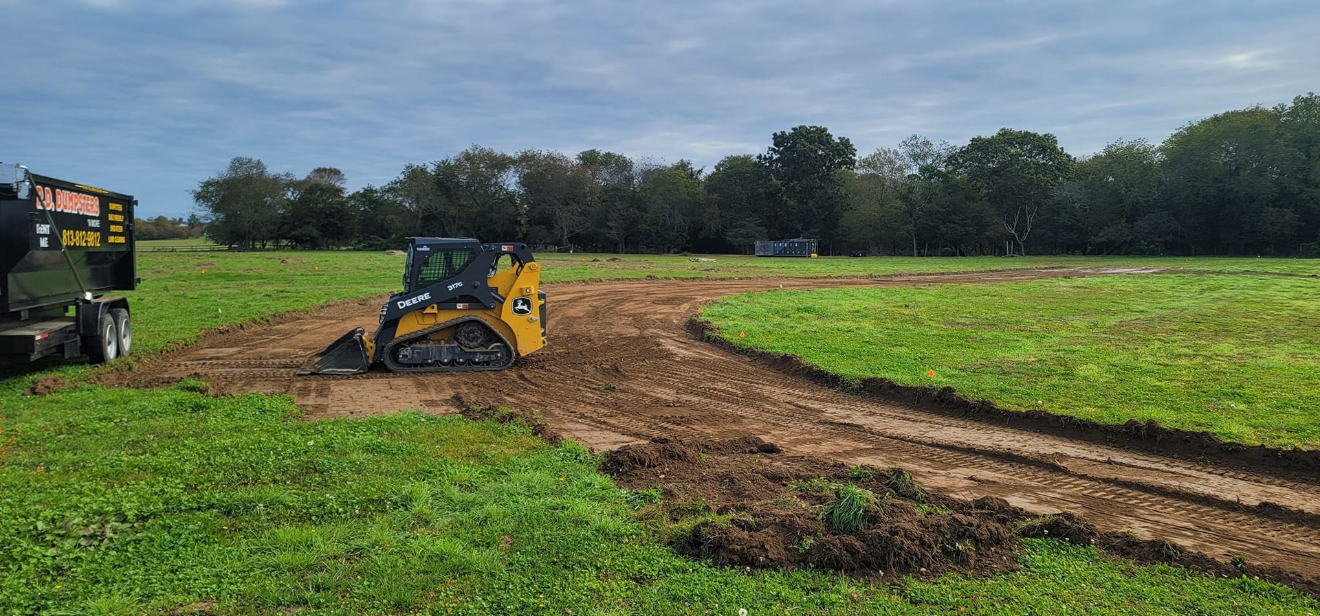 A yellow compact track loader sits on a muddy, unfinished dirt path in a grassy field under a cloudy sky.
