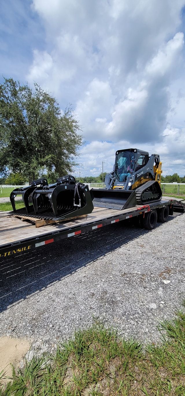 A yellow and black compact track loader and a separate black grapple attachment sit on a flatbed trailer outdoors.