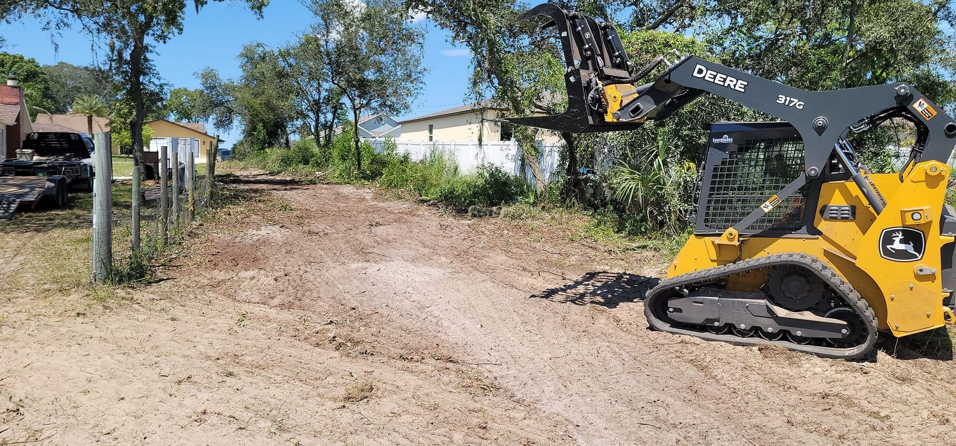 A yellow tracked skid steer with a grapple attachment parked on a dirt lot in a rural area with trees and buildings.