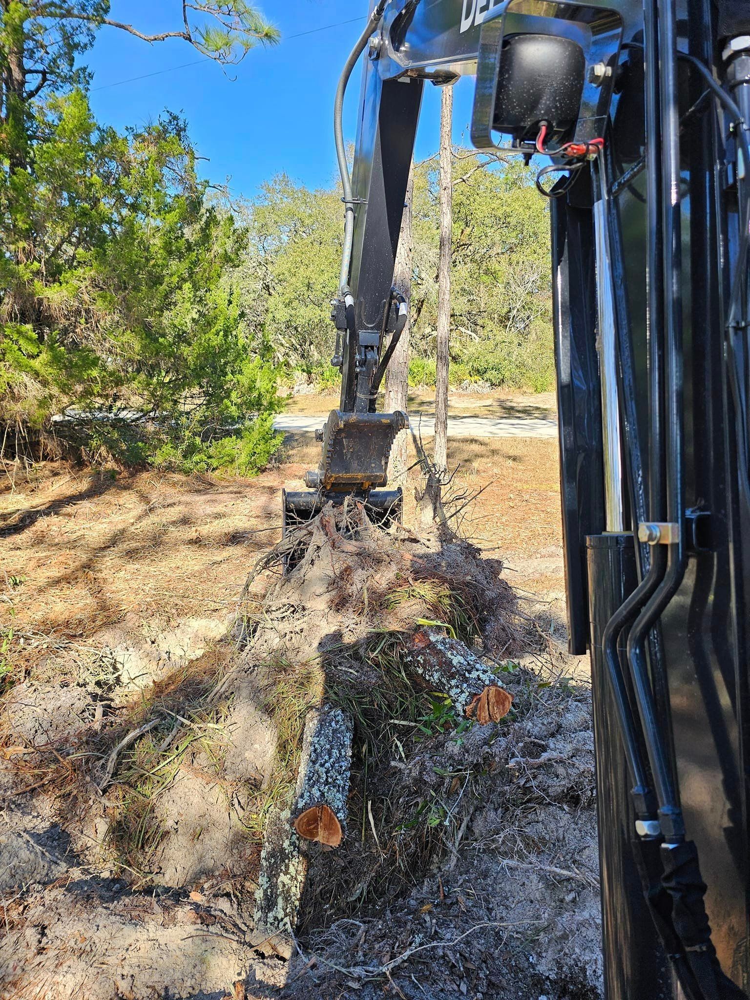 An excavator arm with a grapple attachment moves fallen logs in a sunny, wooded landscape.
