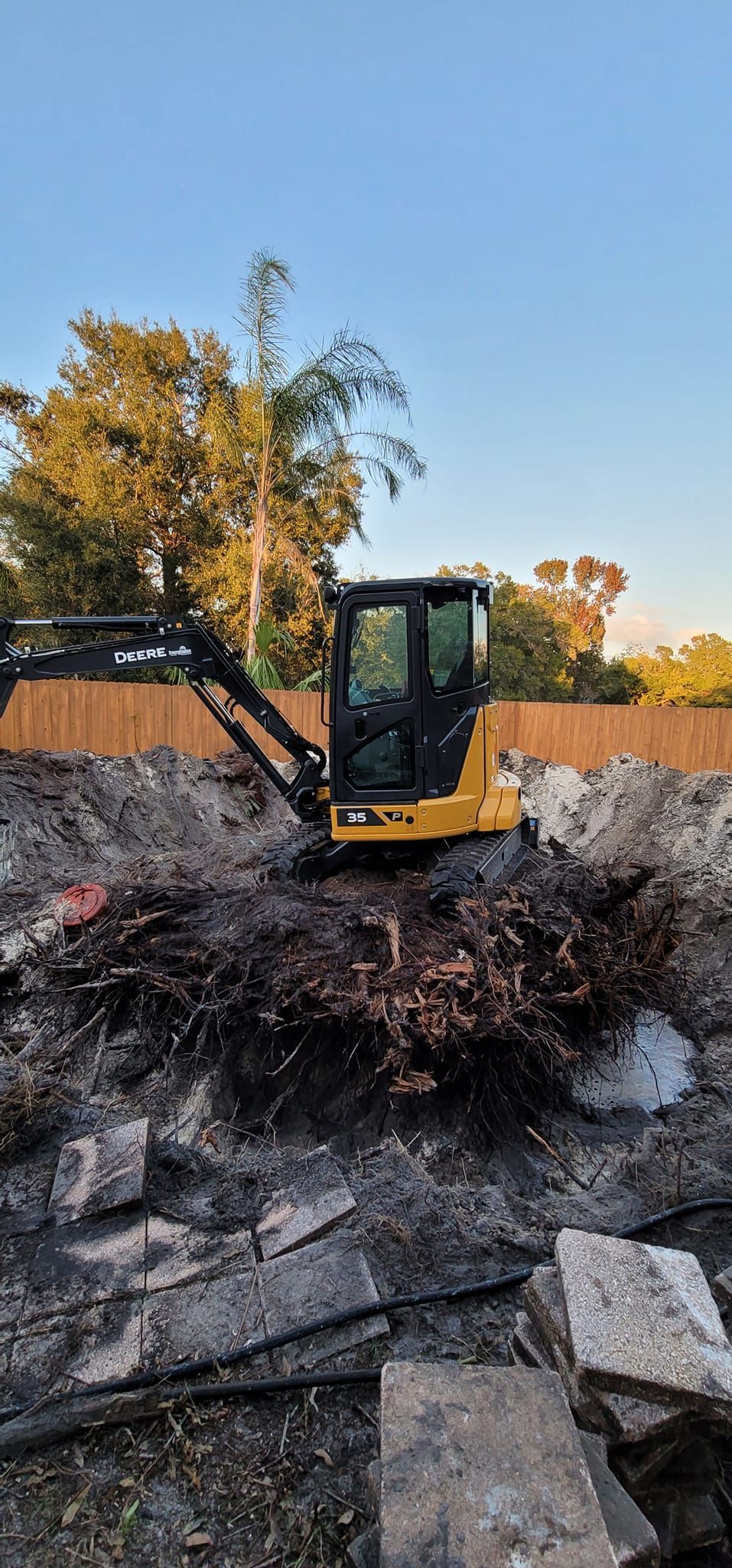 A yellow mini excavator sits in a muddy construction site filled with debris and dark, damp soil under a clear blue sky.