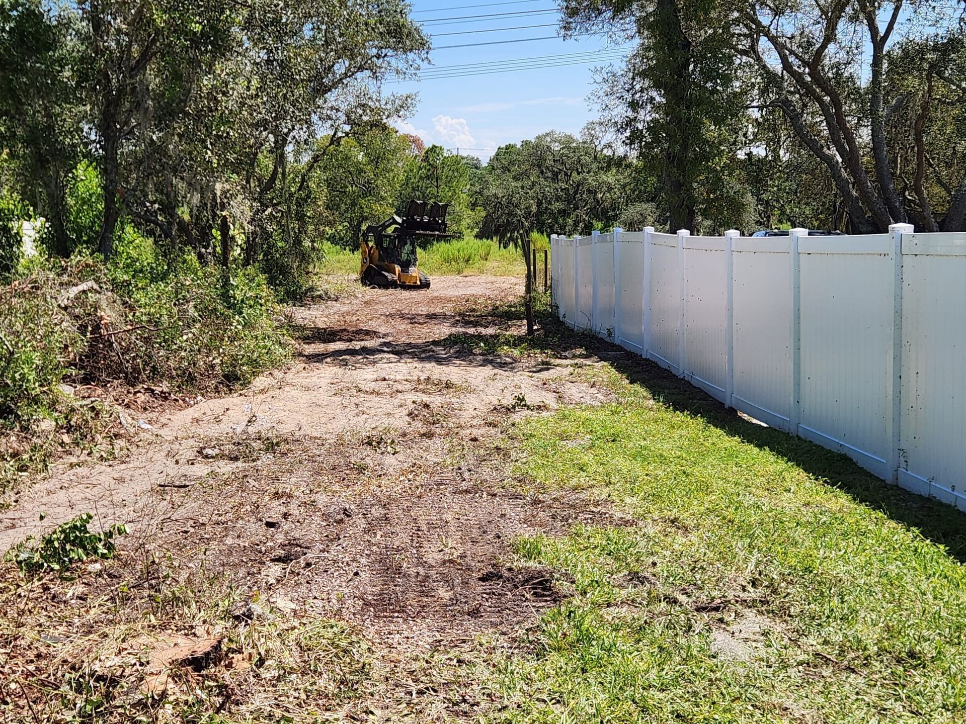 A construction skid steer clears a wooded area next to a white privacy fence on a sunny day.