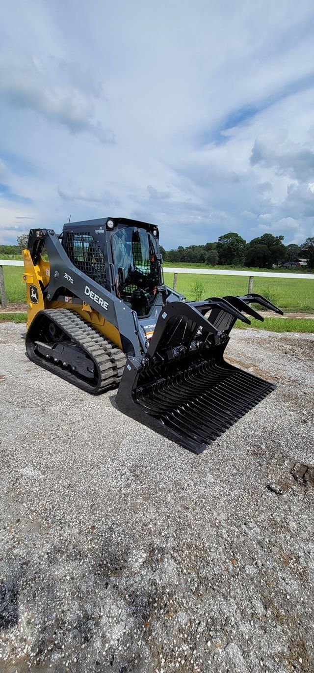 A yellow and dark gray John Deere compact track loader equipped with a grapple bucket parked on a gravel lot.