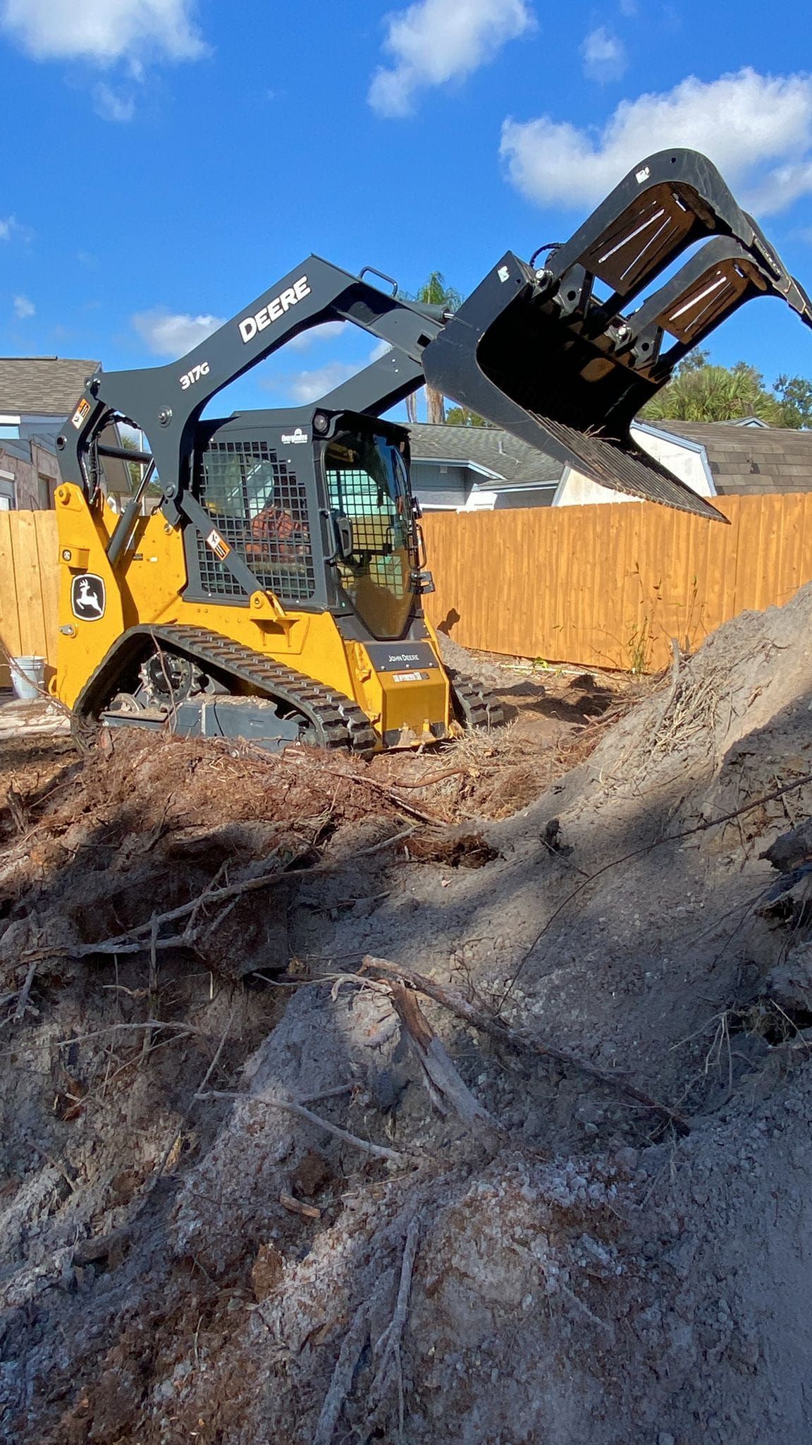 A yellow John Deere skid steer with a grapple bucket works on a dirt construction site in front of a wooden fence.