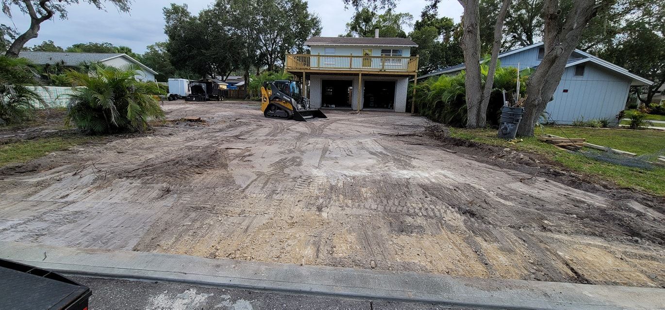 A yellow skid steer sits on a cleared, dirt residential driveway in front of a two-story house with a garage.