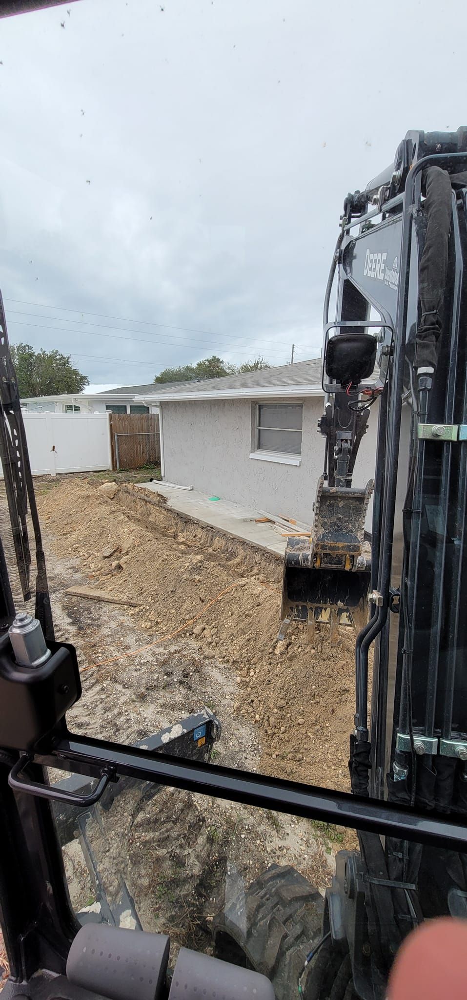 View from inside the cab of an excavator looking out at a residential construction site with a light-colored house.