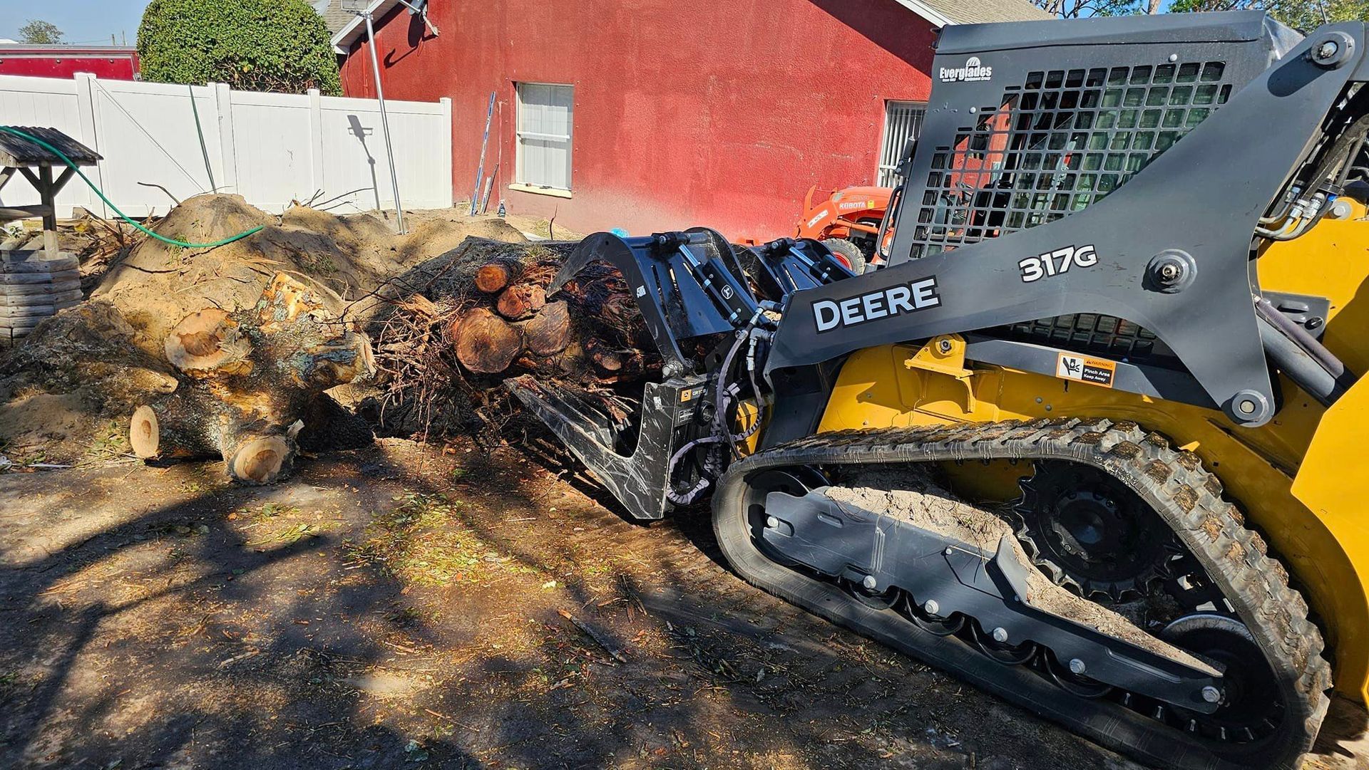 A yellow John Deere compact track loader with a grapple attachment moves a pile of tree logs and debris in a yard.