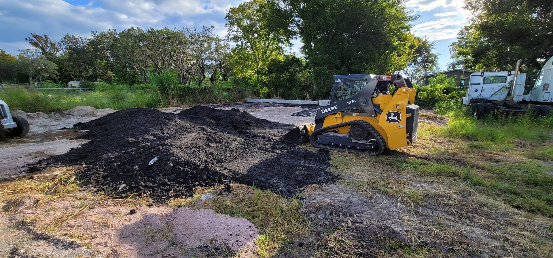 A yellow skid-steer loader sits in a clearing with a pile of dark gravel, surrounded by trees and vegetation.