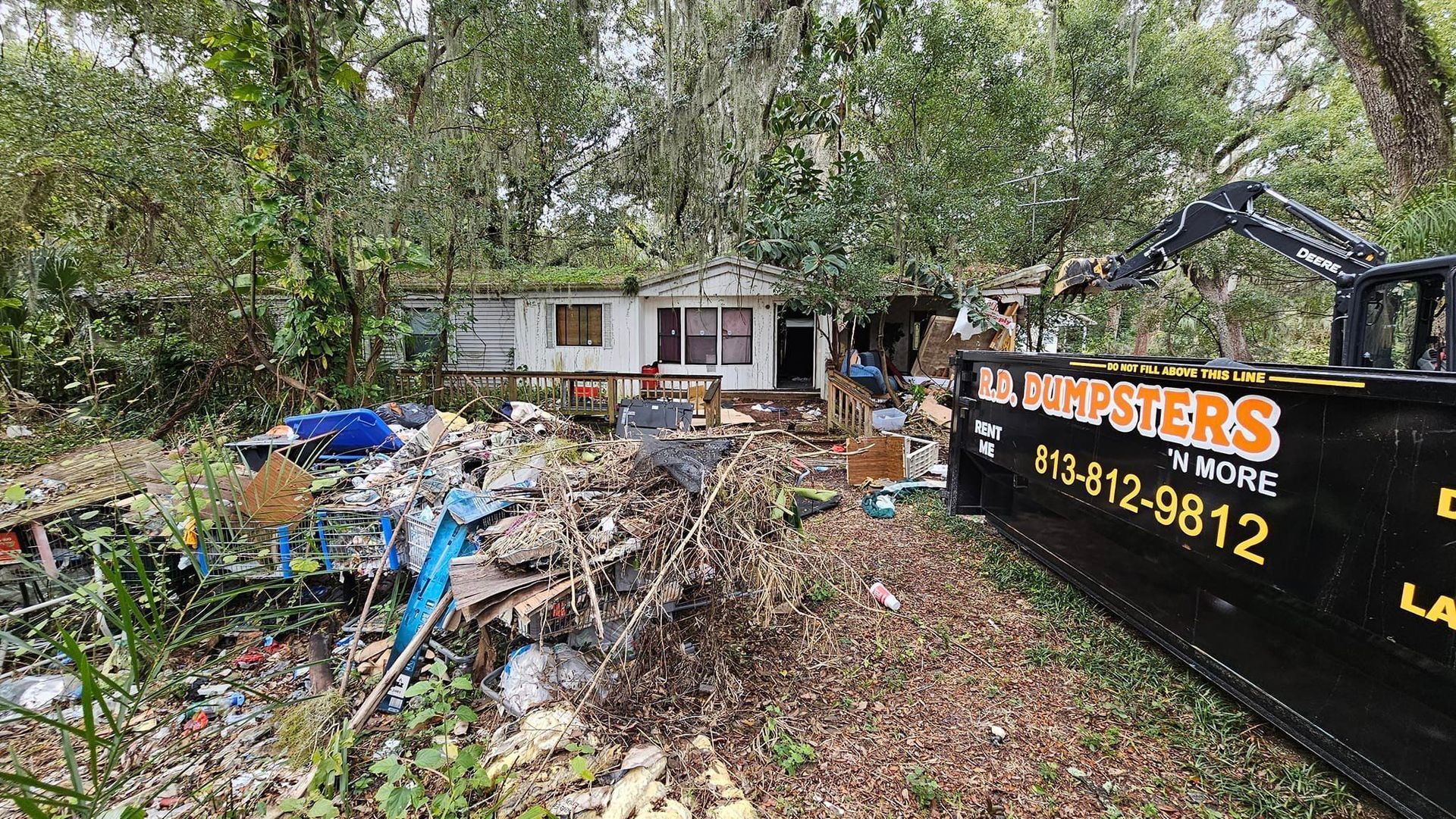 A house cluttered with debris sits in a wooded lot next to a black dumpster labeled 
