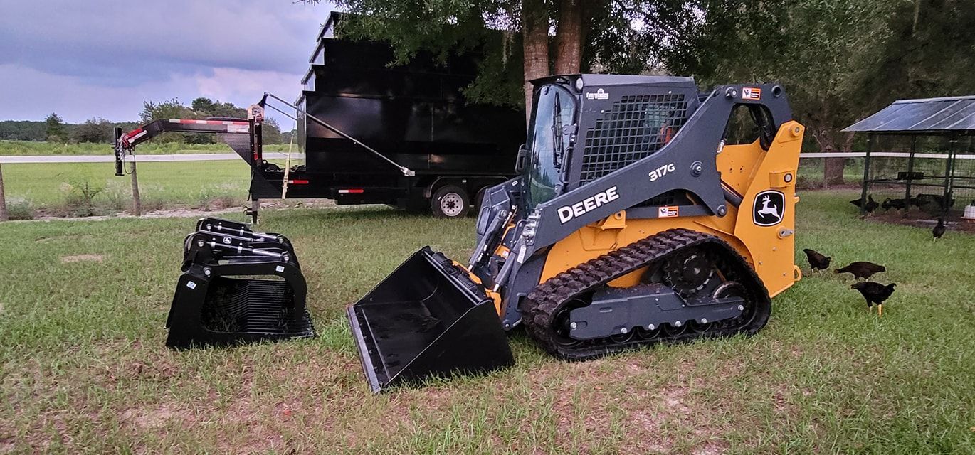 A yellow and black John Deere track loader parked on a grassy field next to an attachment, with a trailer in the distance.