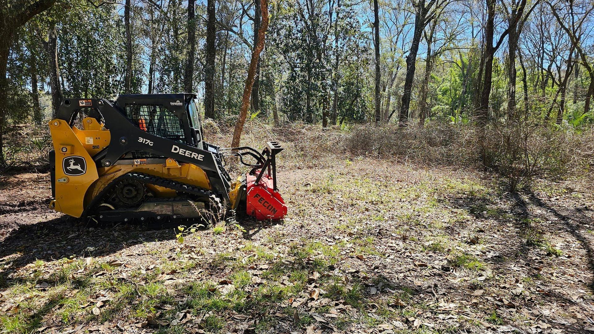 A yellow John Deere compact track loader with a red forestry mulcher attachment sits in a sunlit, wooded area.