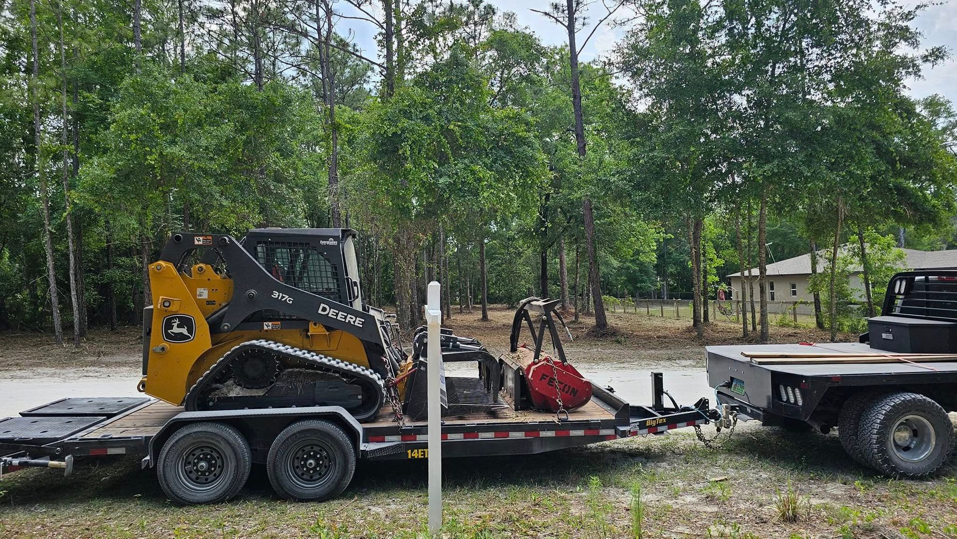 A yellow John Deere compact track loader with a red attachment on a flatbed trailer being towed on a rural dirt road.