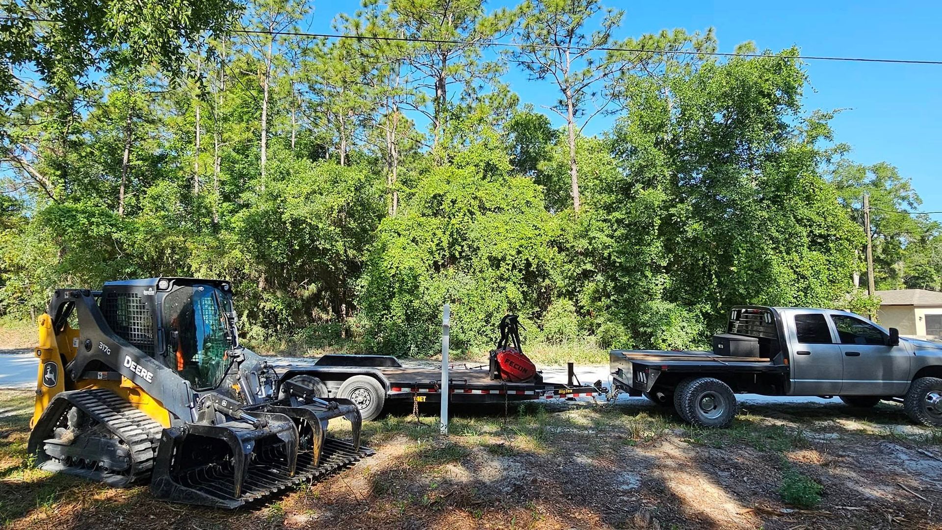 A yellow skid steer loader parked on a dirt lot next to a flatbed trailer and a silver pickup truck by a treeline.