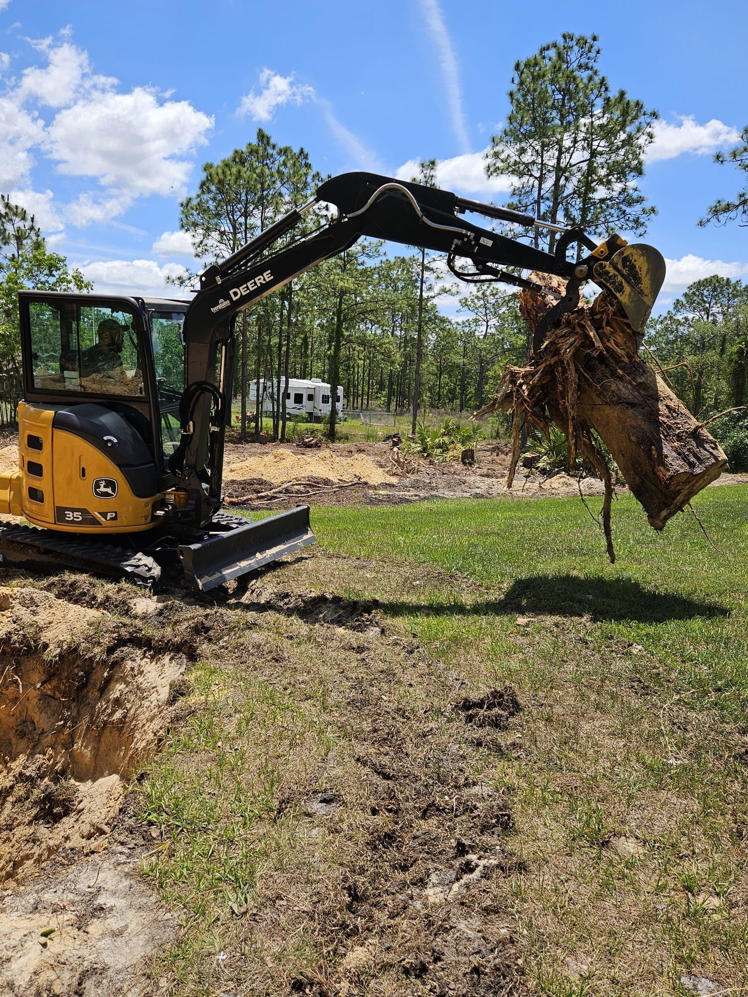 A yellow mini excavator lifts a large tree stump from a hole in a grassy, sunlit field.