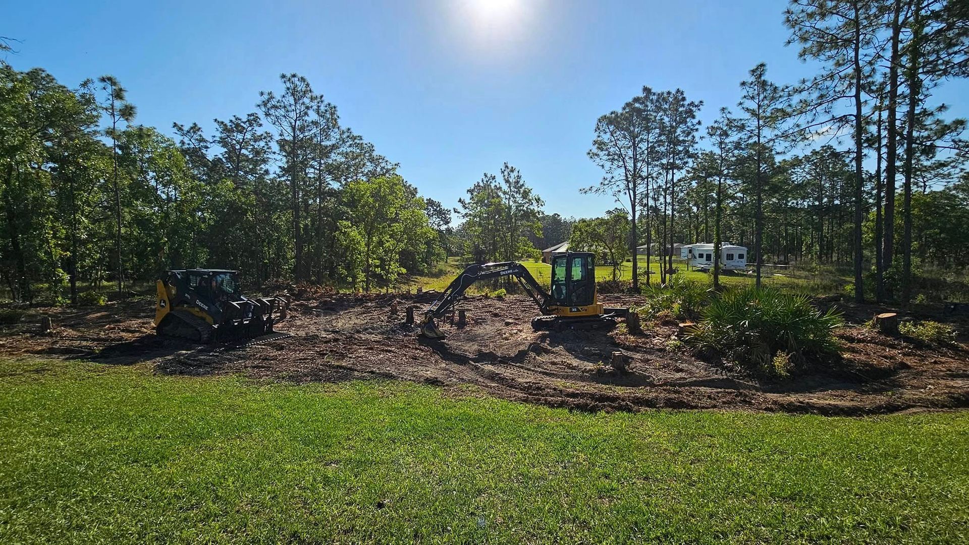 An excavator and a skid steer clear trees and brush in a sunny, wooded lot.