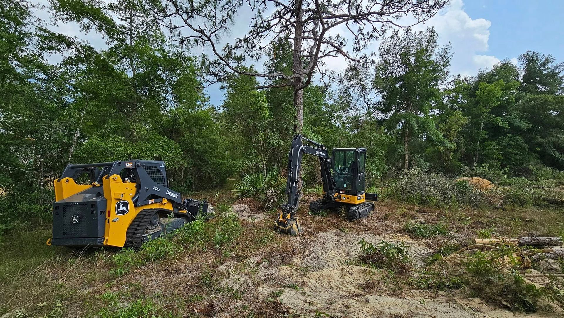 A yellow skid steer and a small excavator working to clear trees and brush in a wooded area on a sunny day.