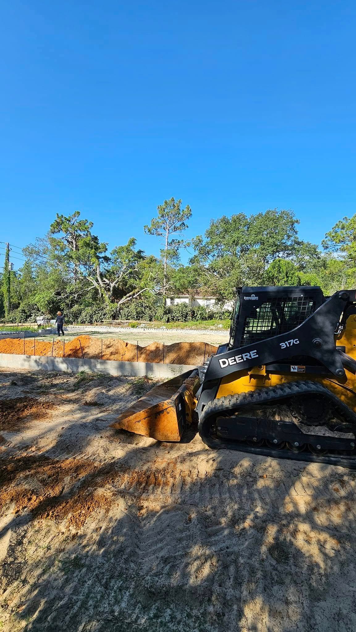 A yellow skid-steer loader parked on a sandy construction site with a green forest in the background under a blue sky.