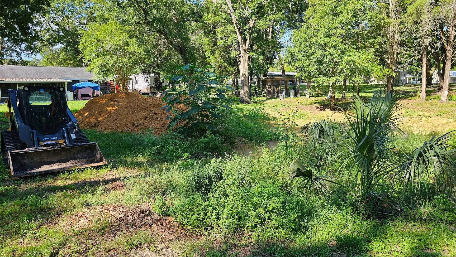 A blue skid steer parked next to a large pile of dirt in a sunny, tree-lined, grassy lot.