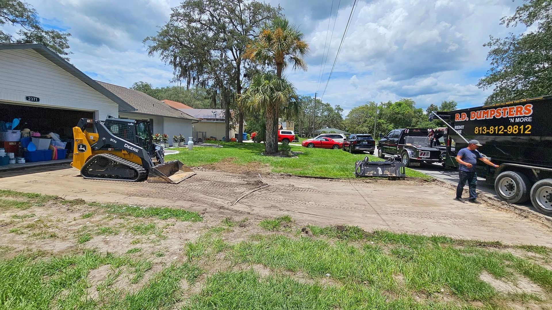 A person uses a yellow skid-steer loader to clear debris from a residential yard into a dump trailer.