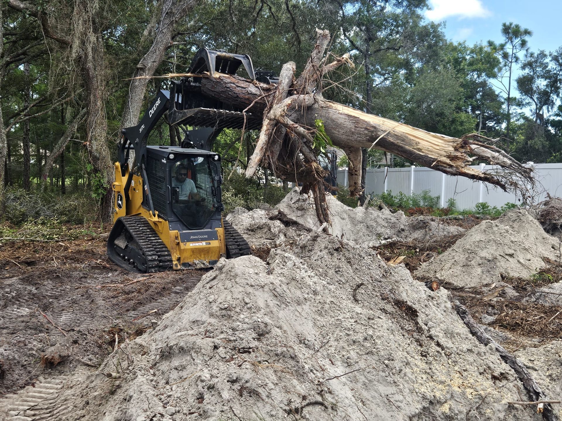 A yellow skid-steer loader lifts a large tree root ball and trunk at a construction site with piles of dirt.