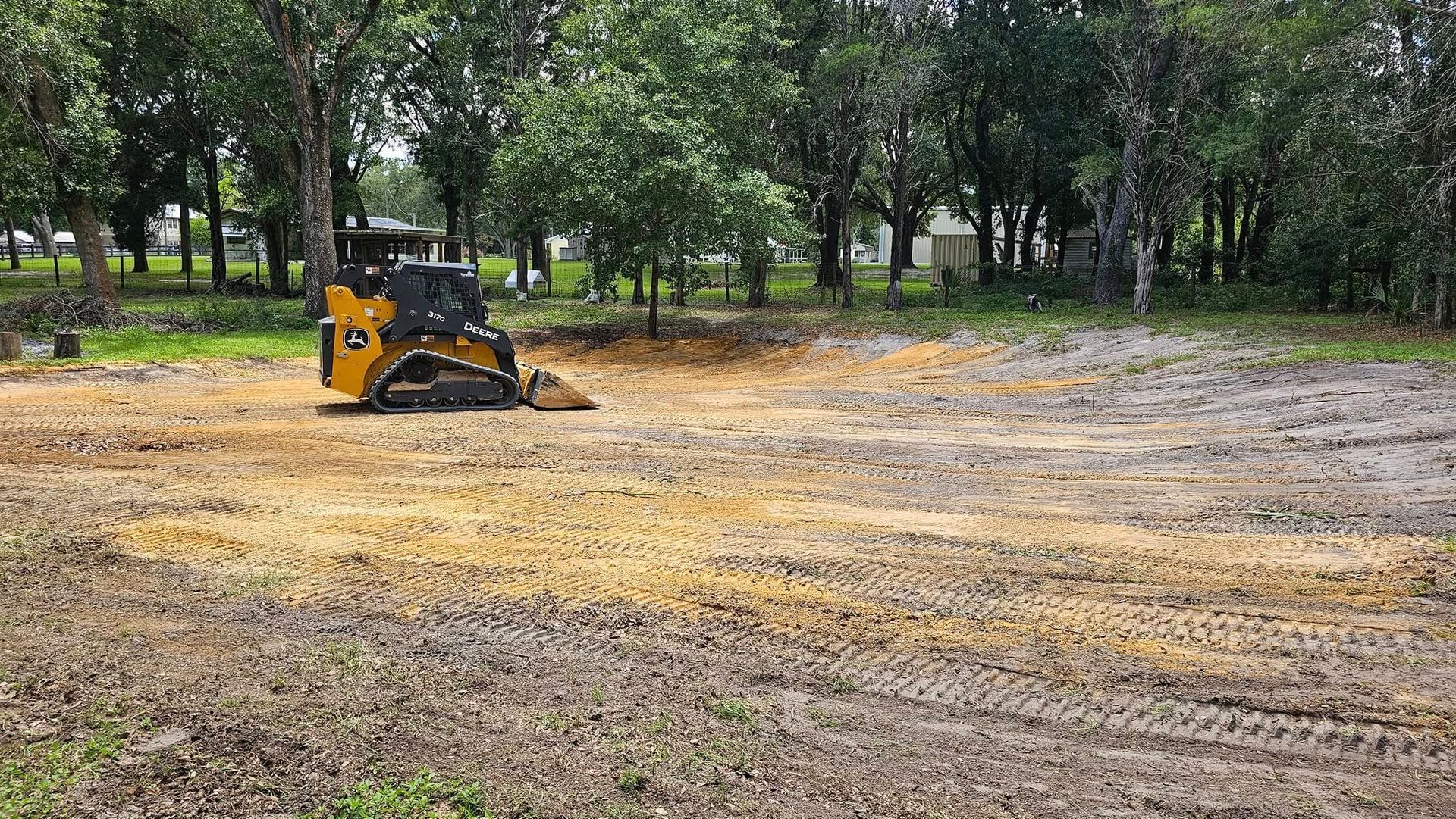 A yellow compact track loader sits on a dirt lot undergoing clearing, with mature trees in the background.