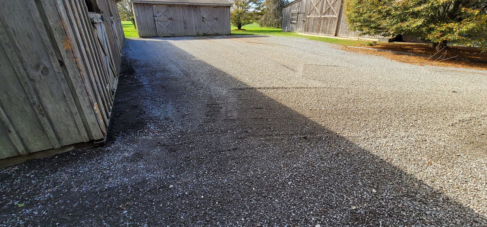 A gravel driveway alongside a wooden building under bright sunlight, leading toward a distant garage.