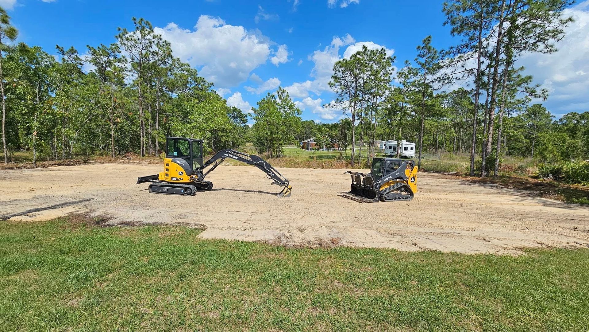 A yellow excavator and a skid steer parked on a cleared sandy lot surrounded by green trees under a blue sky.