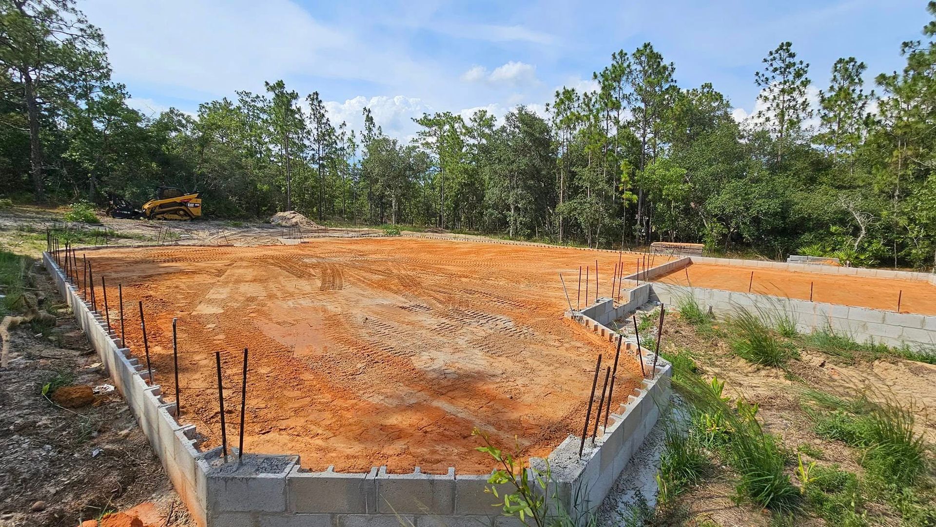 A construction site showing a concrete block foundation for a building filled with red dirt amidst a wooded area.