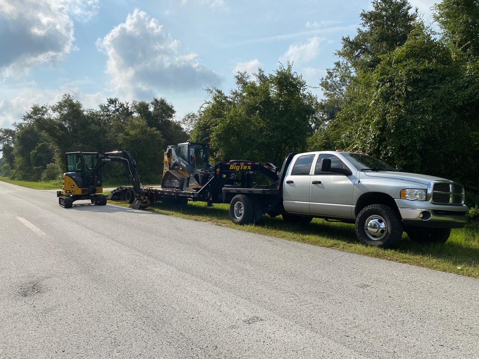 A silver pickup truck towing a flatbed trailer with a small excavator on it, parked on the side of a rural road.