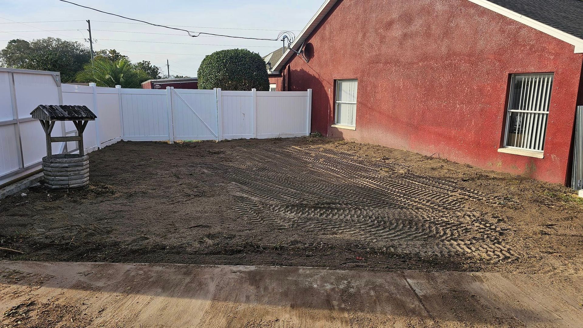 A fenced backyard with freshly tilled soil, a stone wishing well, and a red brick house with two windows.