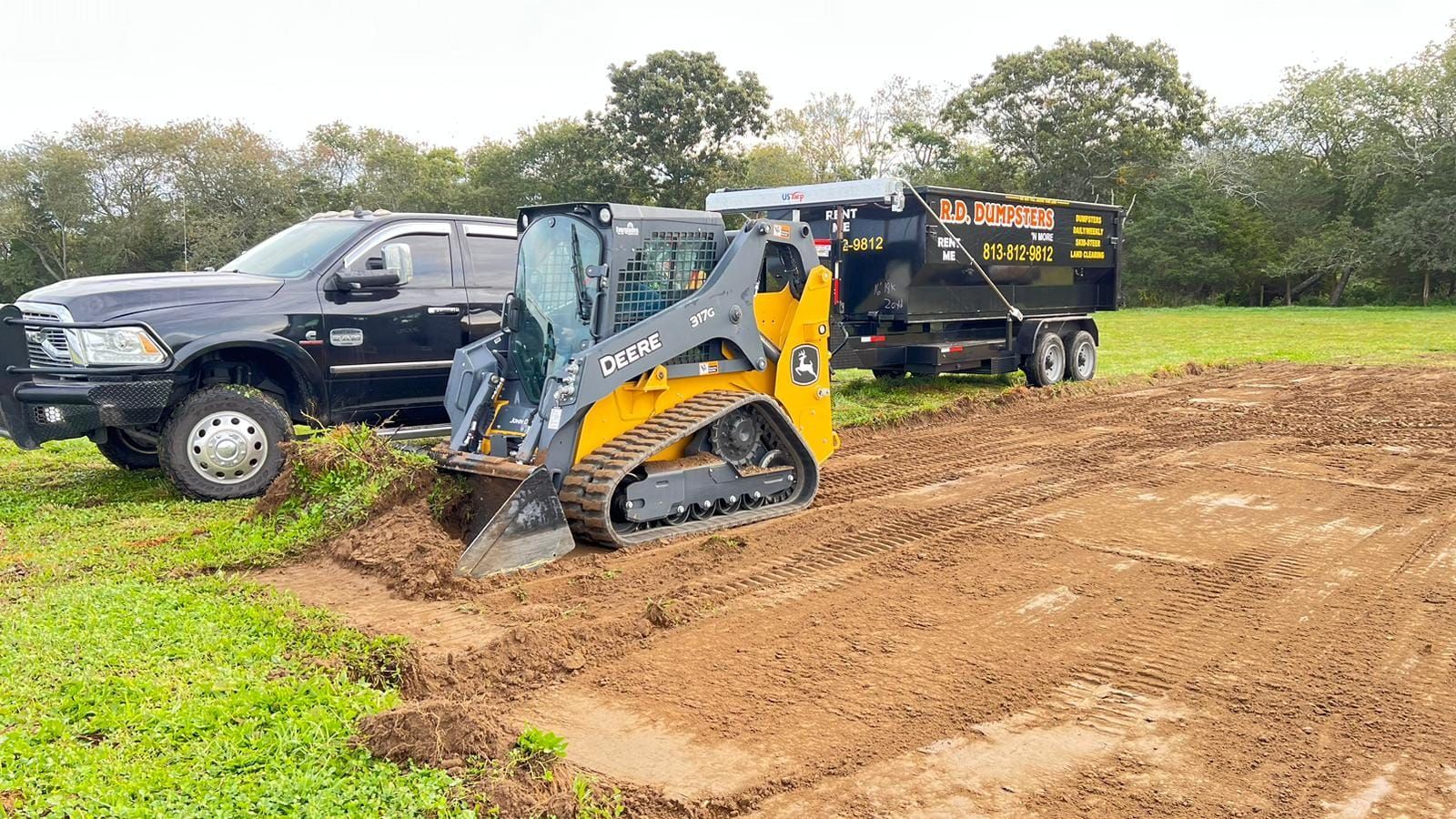 A yellow Caterpillar skid steer loader parked next to a black pickup truck and a dumpster trailer in a muddy field.