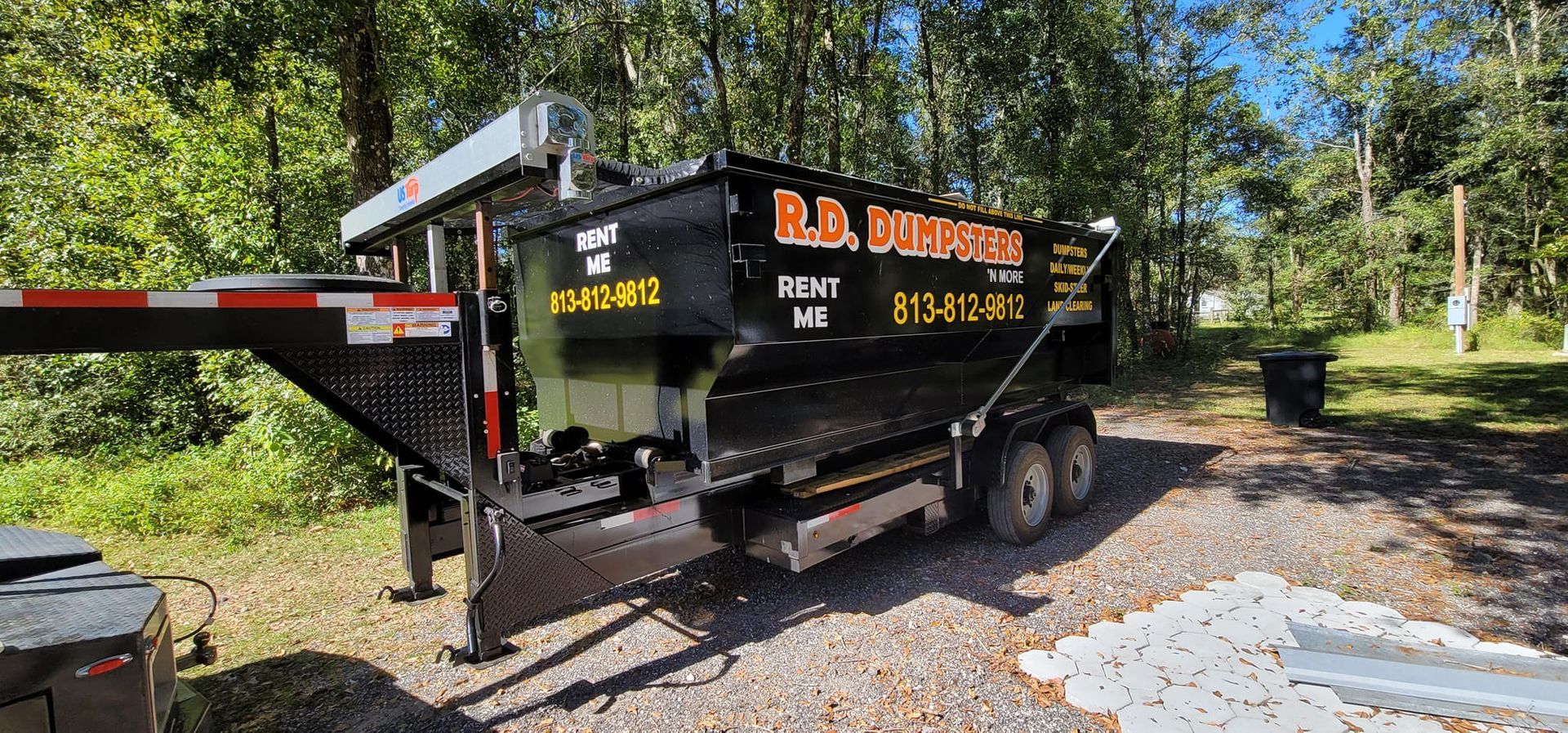 A black dump trailer parked on a gravel surface outdoors with trees in the background.
