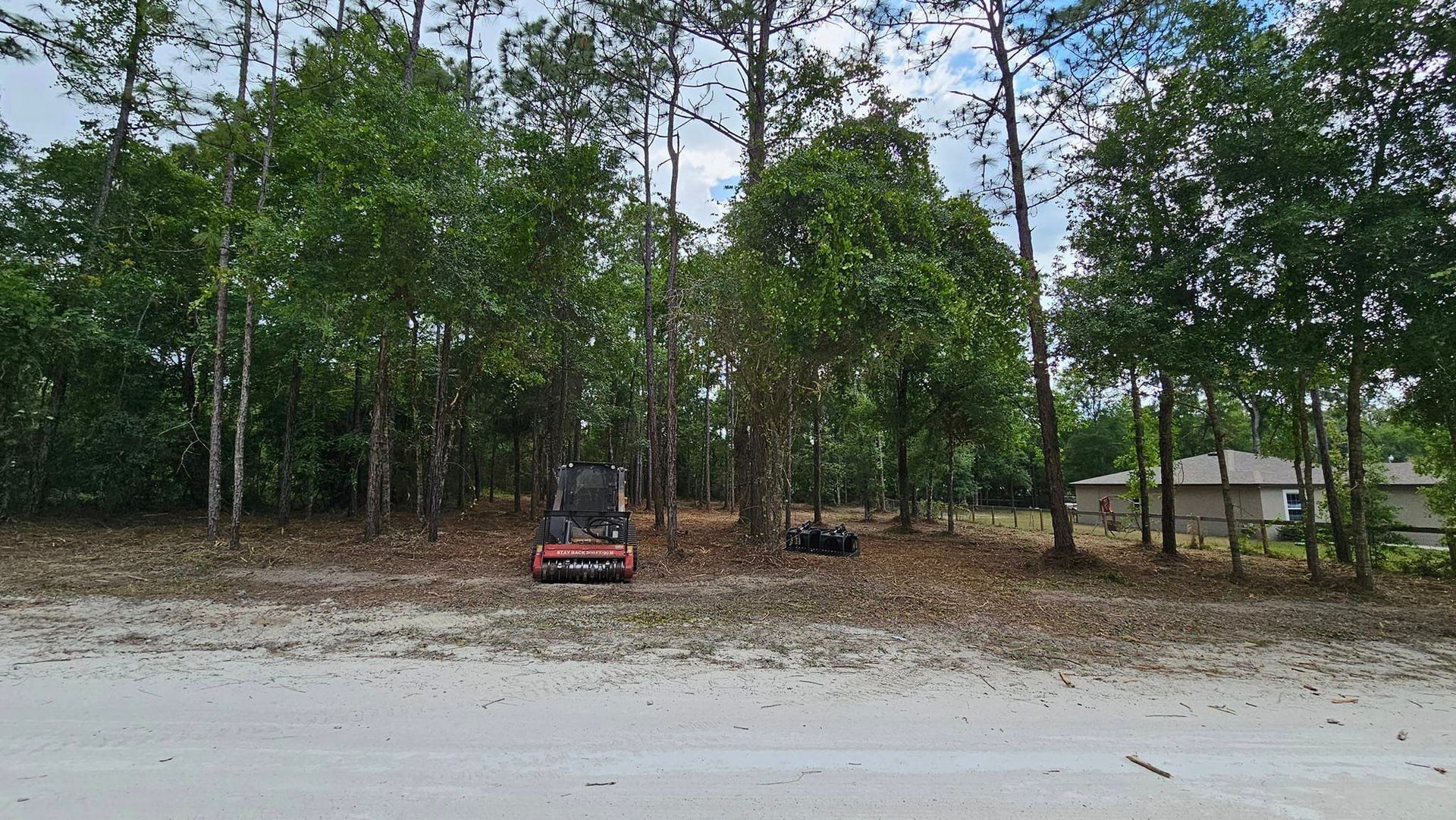 A red skid steer loader sits on a sandy, wooded lot with trees and a house visible in the background.