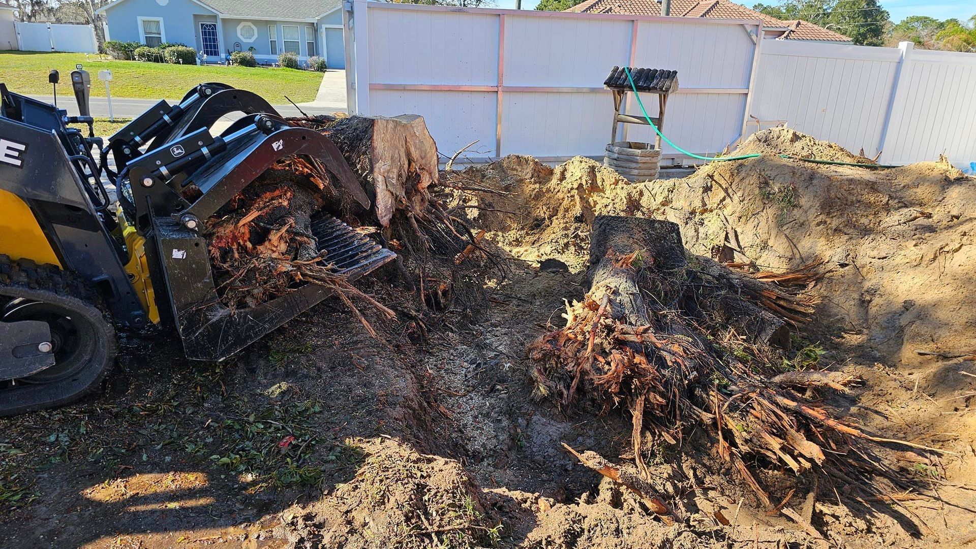 A skid steer with a grapple attachment moves a large, unearthed tree stump in a residential yard.