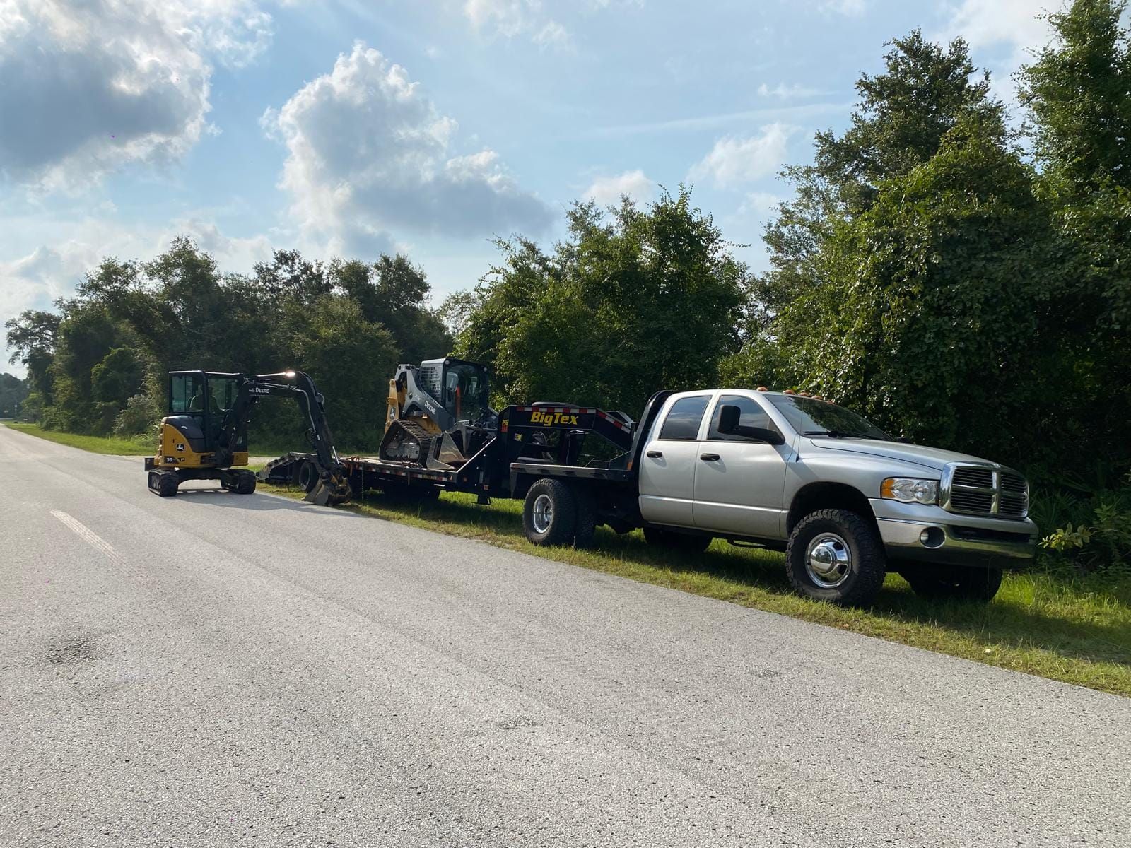 A silver pickup truck pulls a flatbed trailer loaded with a skid steer and a small excavator parked on a grassy roadside.