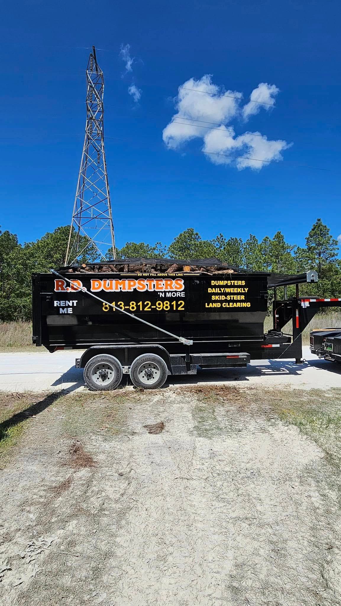 A black dumpster rental trailer sits on a gravel road in front of a tall utility tower under a clear blue sky.