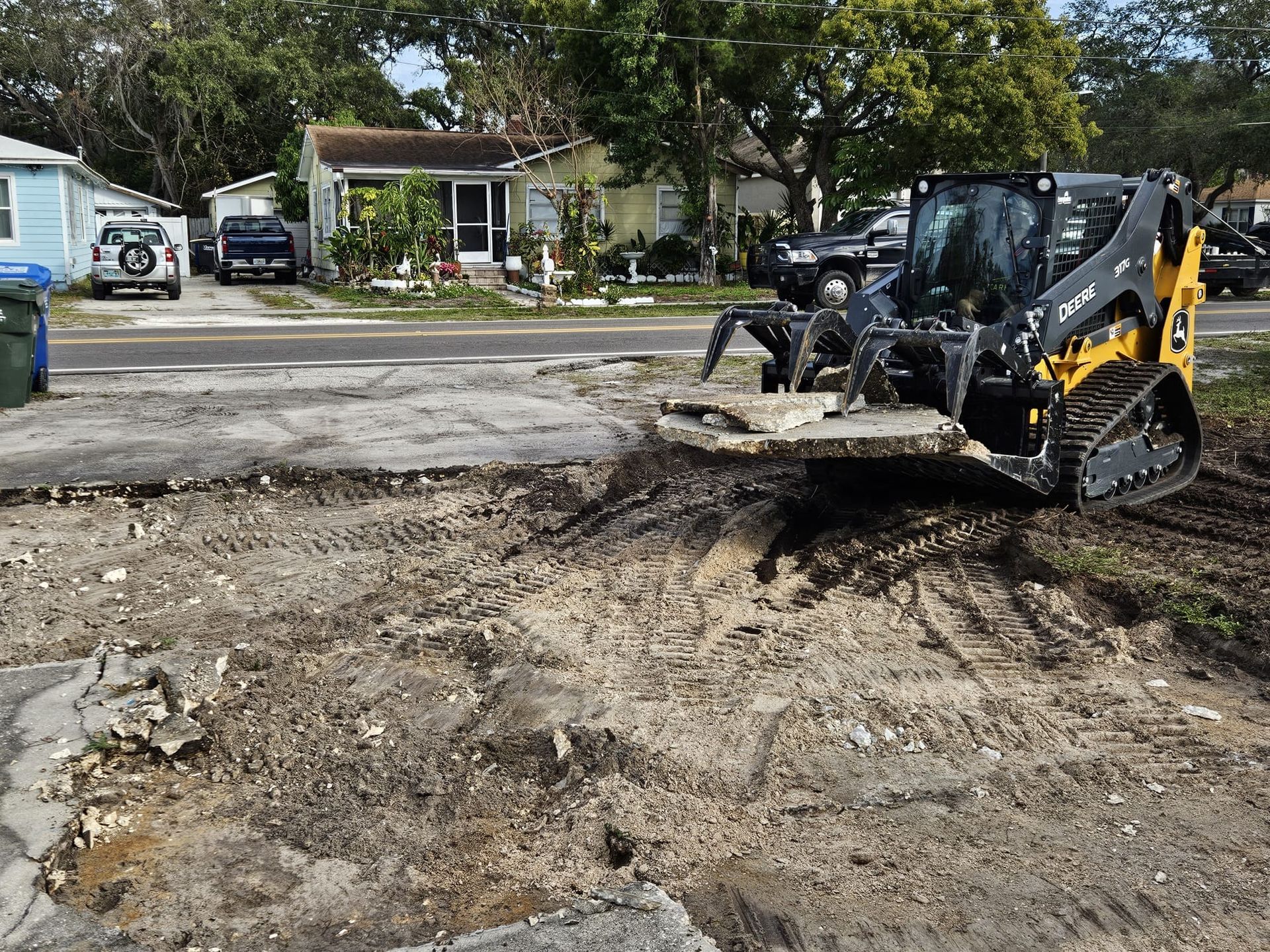 A yellow and black skid-steer loader lifts a large slab of concrete from a dirt construction site in a residential area.
