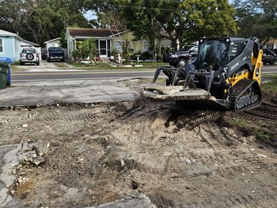 A yellow and black skid-steer loader lifts a large slab of broken concrete from a dirt construction site.