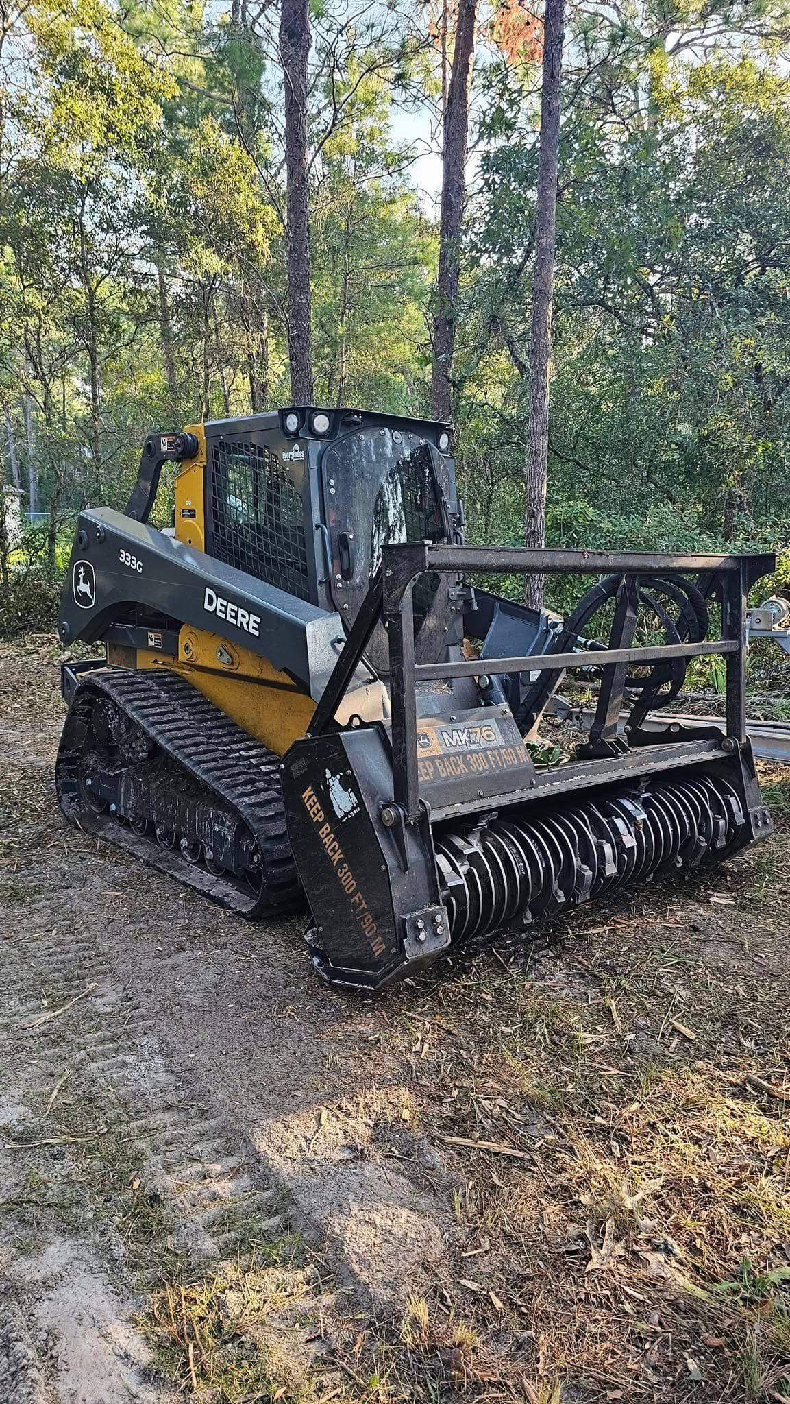 A yellow and black skid-steer loader with a heavy-duty brush cutter attachment parked on a leaf-covered forest trail.