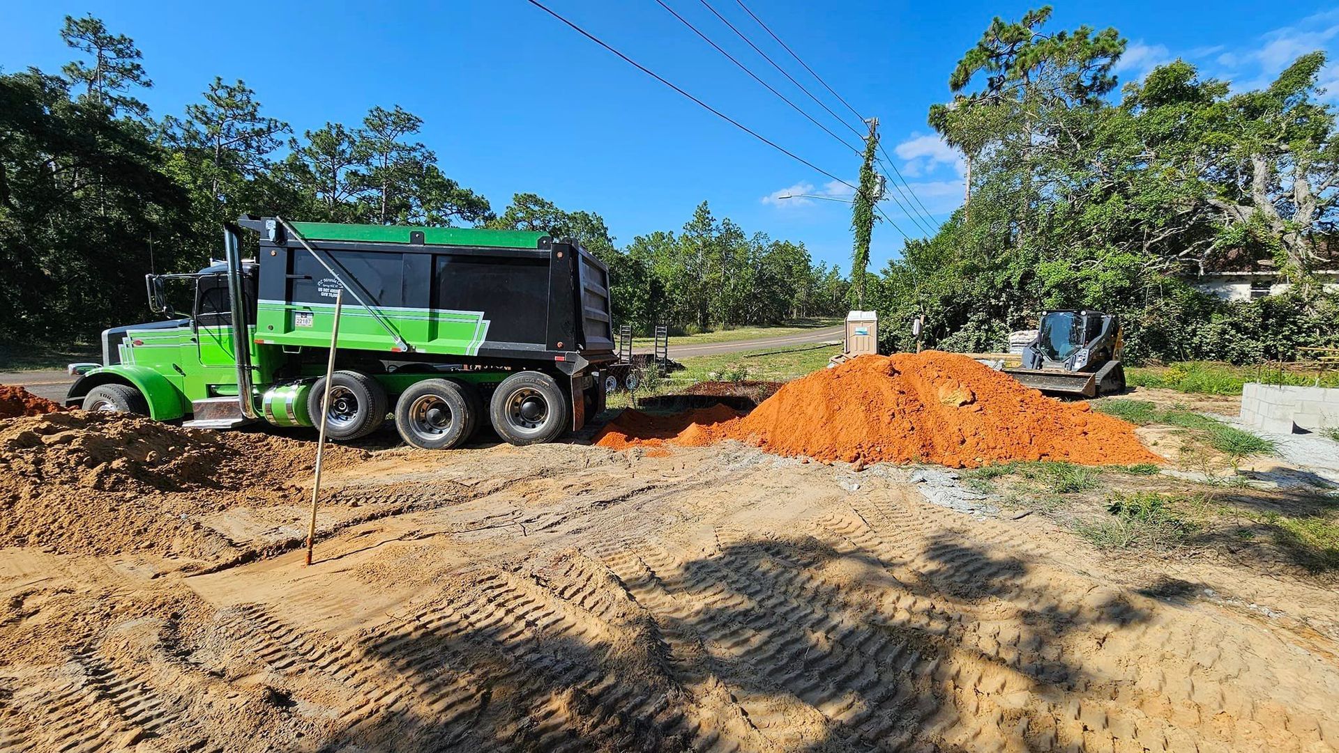 A green dump truck sits next to a large pile of dirt at an outdoor construction site on a sunny day.