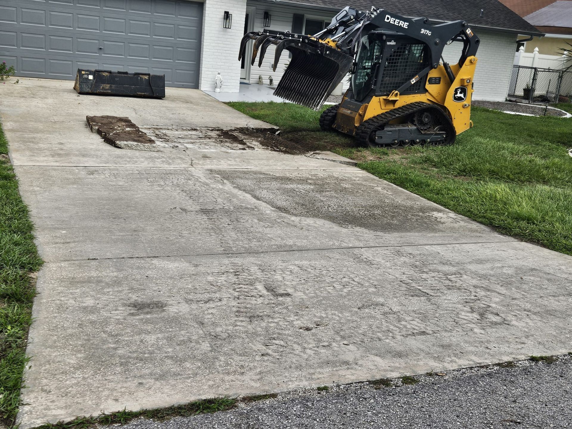 A yellow skid steer with a grapple bucket parked on a driveway near a house, with a section of the concrete removed.