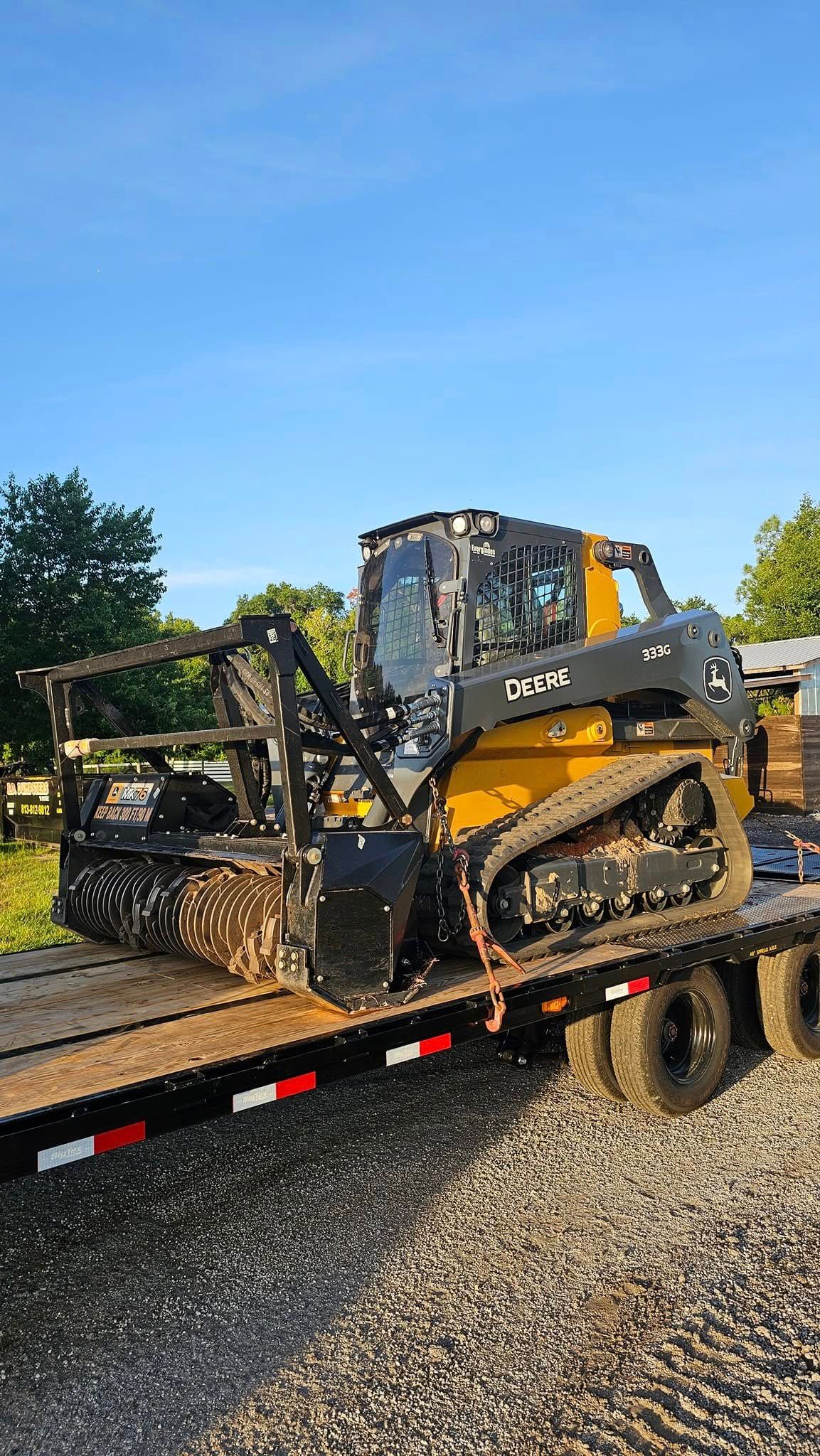 A yellow and black compact track loader with a brush cutter attachment sits on a flatbed trailer outdoors under a blue sky.