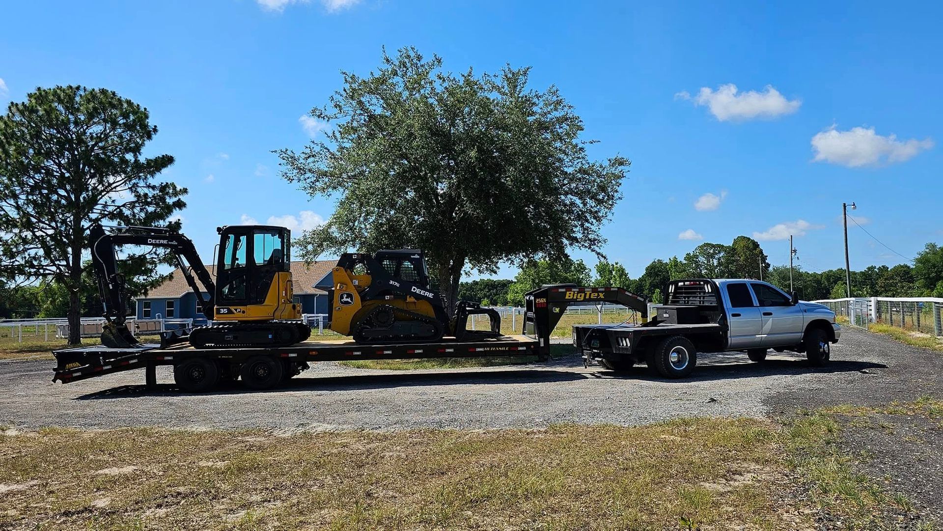 A silver flatbed truck parked on a gravel lot, towing a trailer loaded with a yellow mini-excavator and a skid steer.